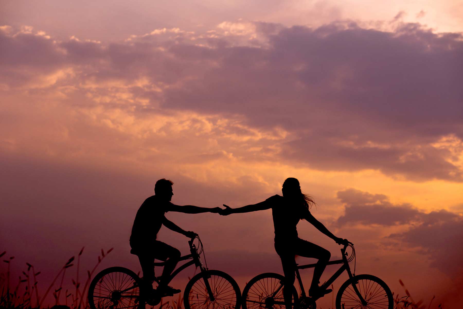 Sunset silhouette of couple on bicycles holding hands.